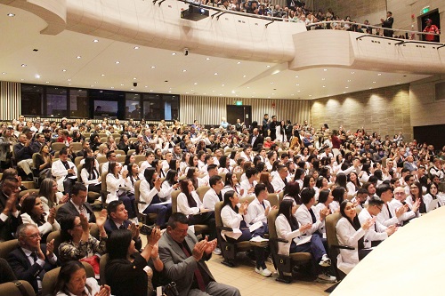 estudiantes de Medicina en bata blanca aplaudiendo durante una ceremonia acompaados por publico en el auditorio