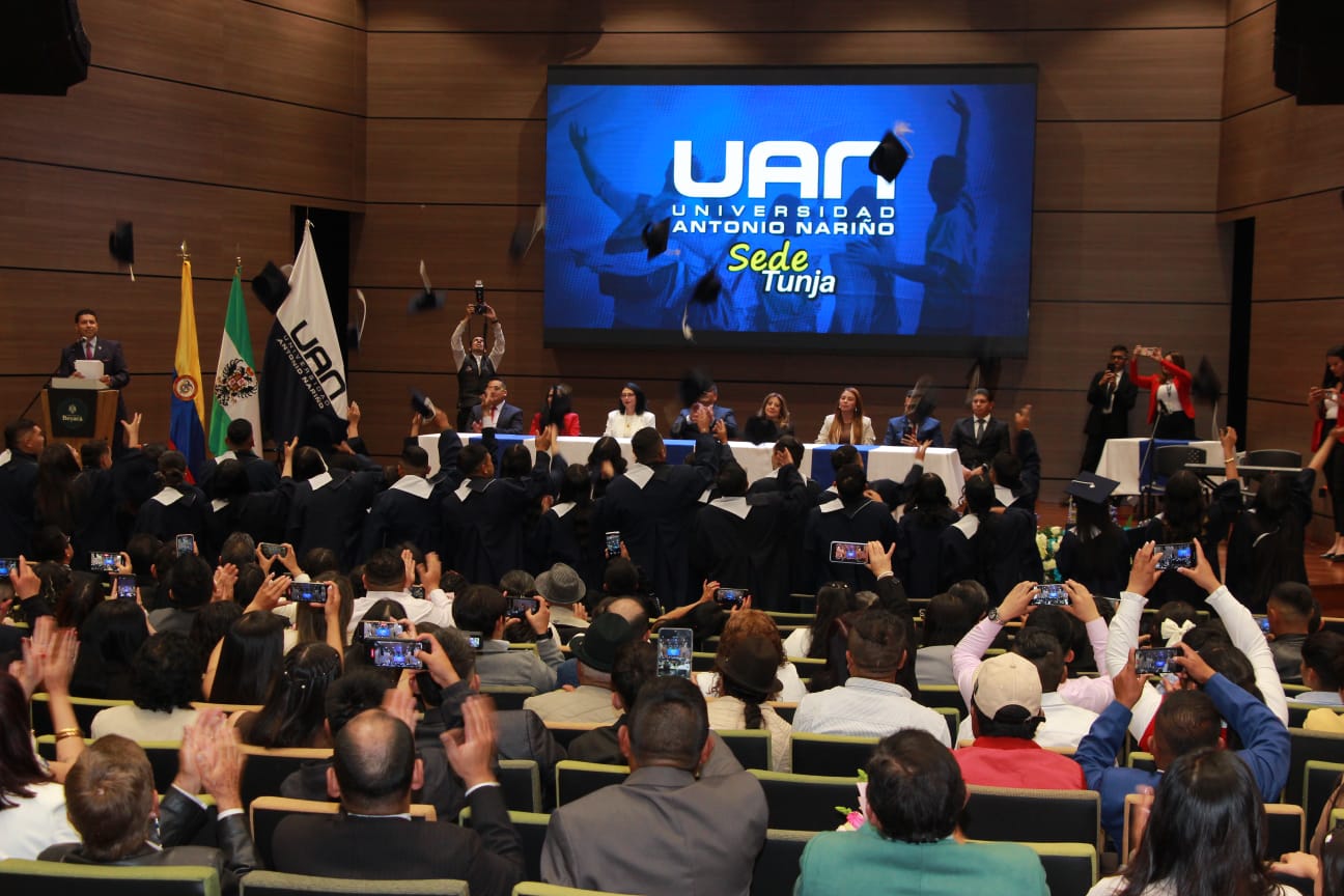 ceremonia de graduacion donde varios estudiantes lanzan sus birretes al aire mientras el publico aplaude y toma fotos En el escenario directivos de la universidad presiden el acto