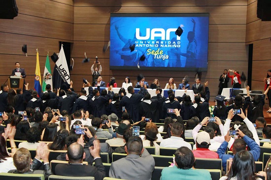 ceremonia de graduacion donde varios estudiantes lanzan sus birretes al aire mientras el publico aplaude y toma fotos En el escenario directivos de la universidad presiden el acto