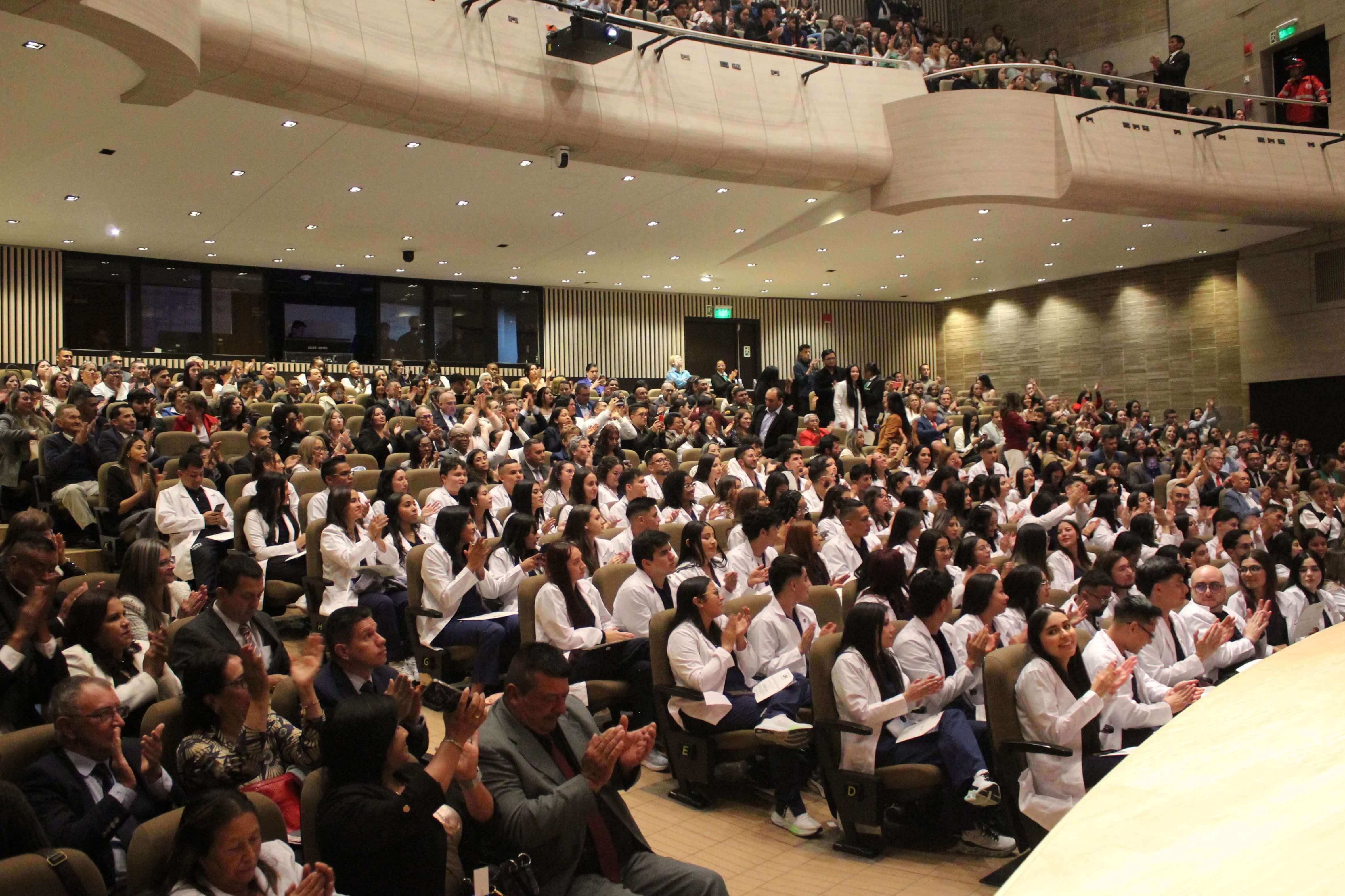 estudiantes de Medicina en bata blanca aplaudiendo durante una ceremonia acompaados por publico en el auditorio