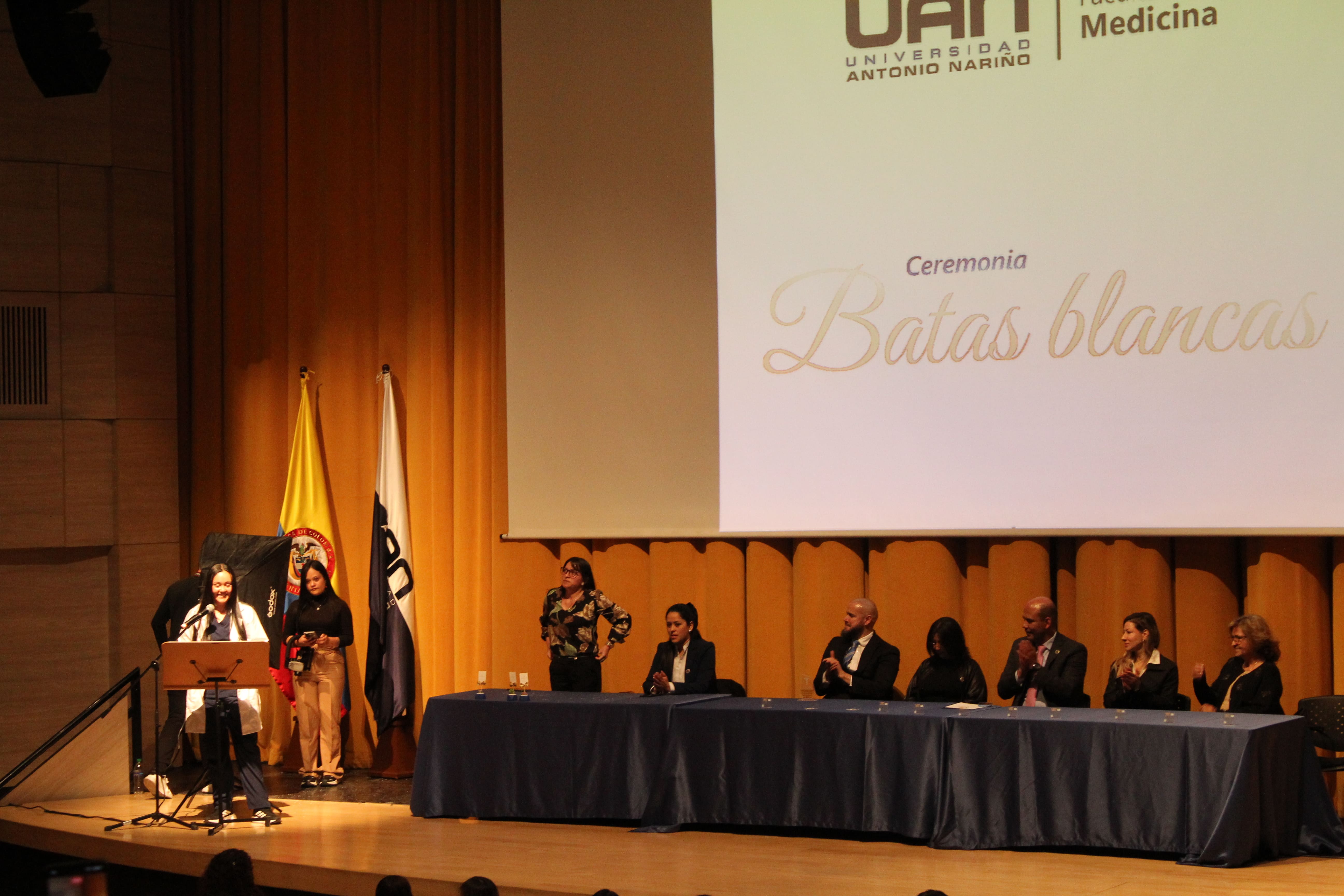 Ceremonia de Batas Blancas de la UAN con una estudiante hablando en el atril mientras el comite academico la acompana desde la mesa principal