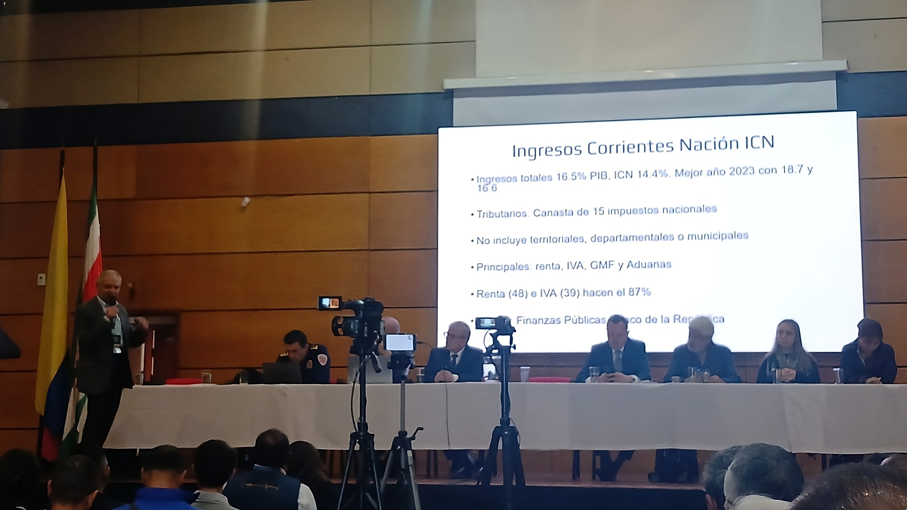 Es una conferencia en un auditorio un panel de personas esta sentado al frente un hombre habla con microfono y una diapositiva grande se proyecta detras mientras el publico escucha