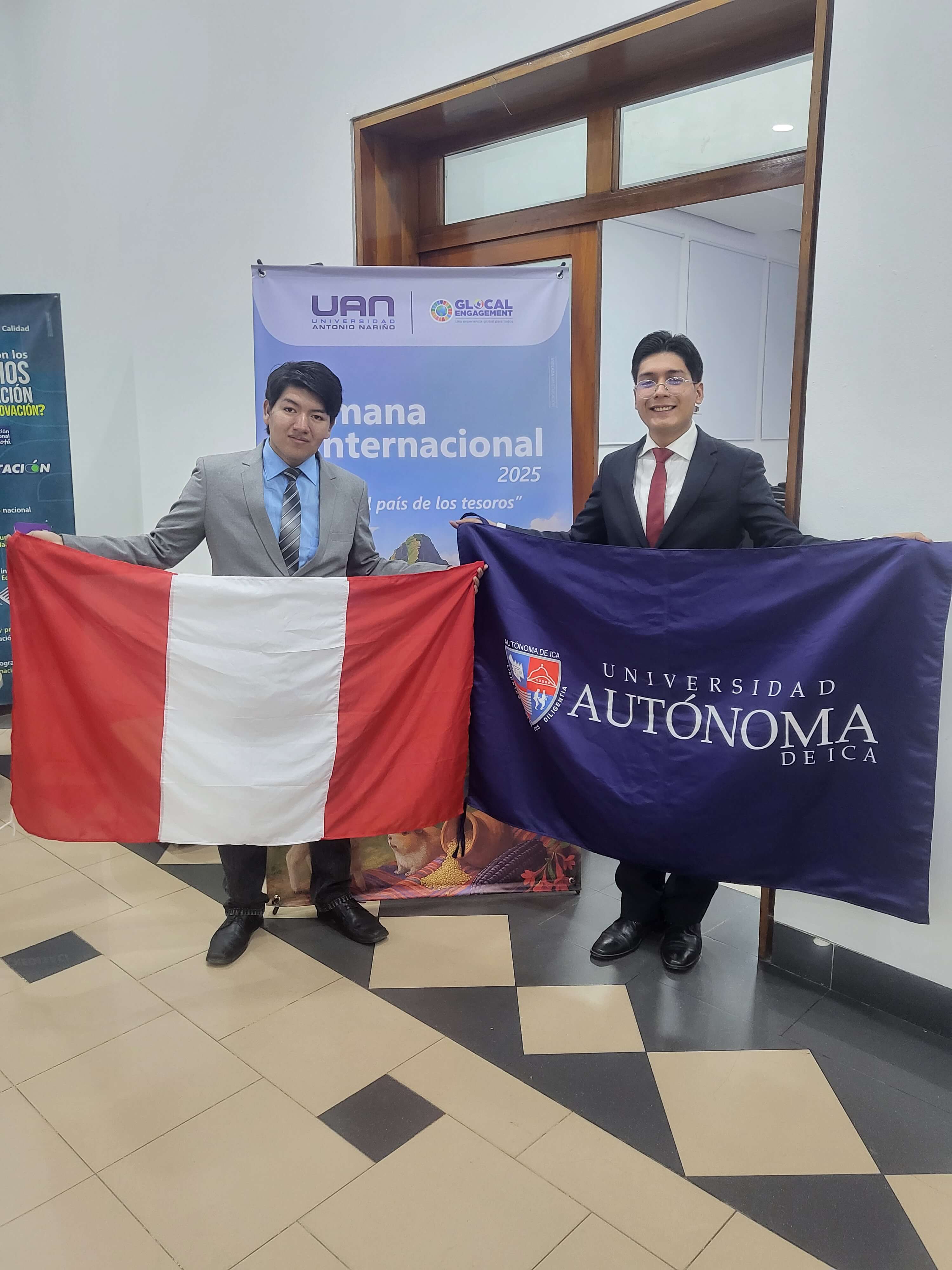 Dos jovenes posan frente a un afiche de la Semana Internacional 2025 Cada uno sostiene una bandera uno la de Peru y el otro la de su universidad, mientras sonrien a la camara