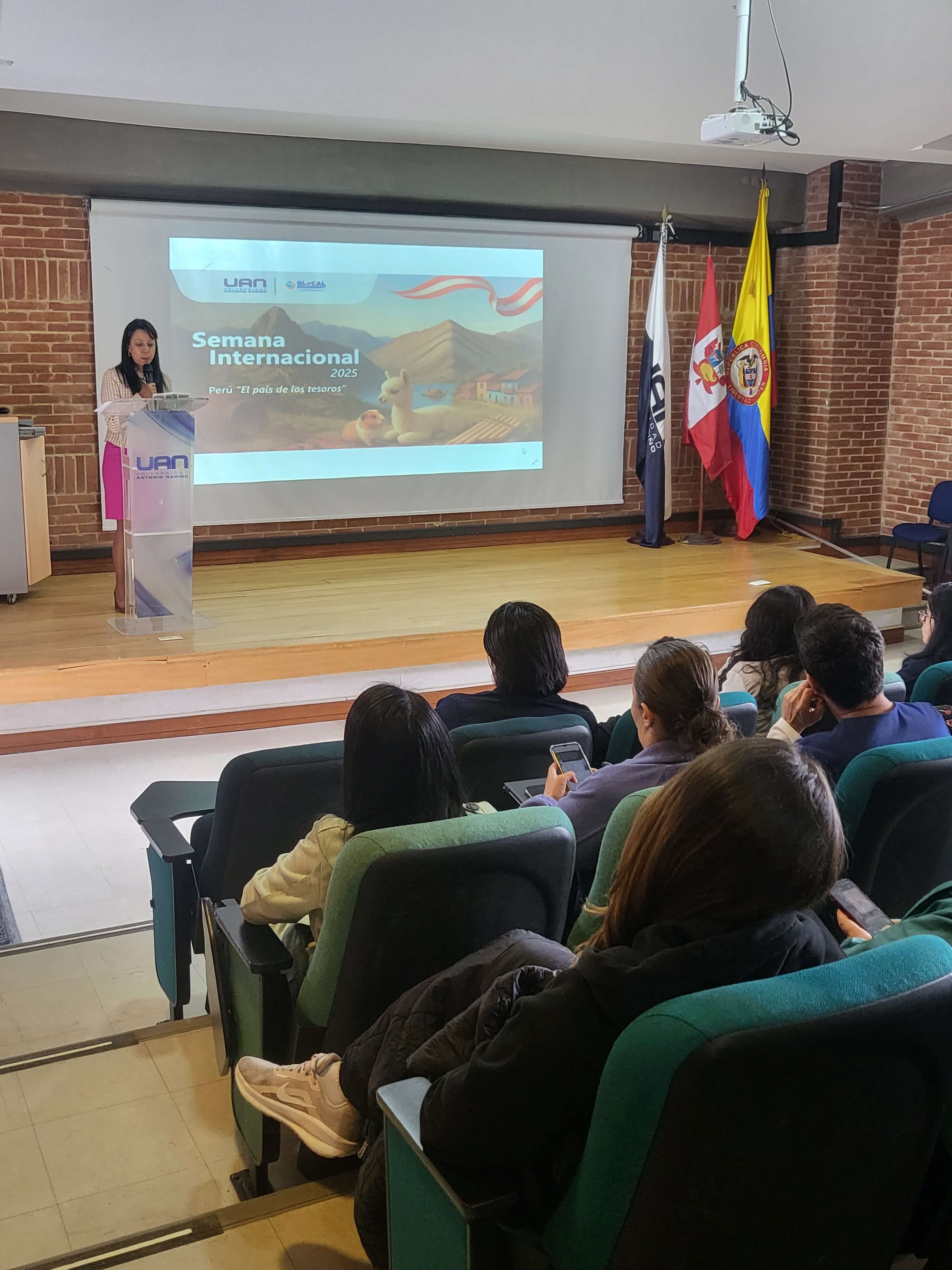 Una mujer habla en un auditorio mientras presenta una diapositiva sobre la Semana Internacional 2025  Peru El país de los tesoros ante un grupo de asistentes sentados