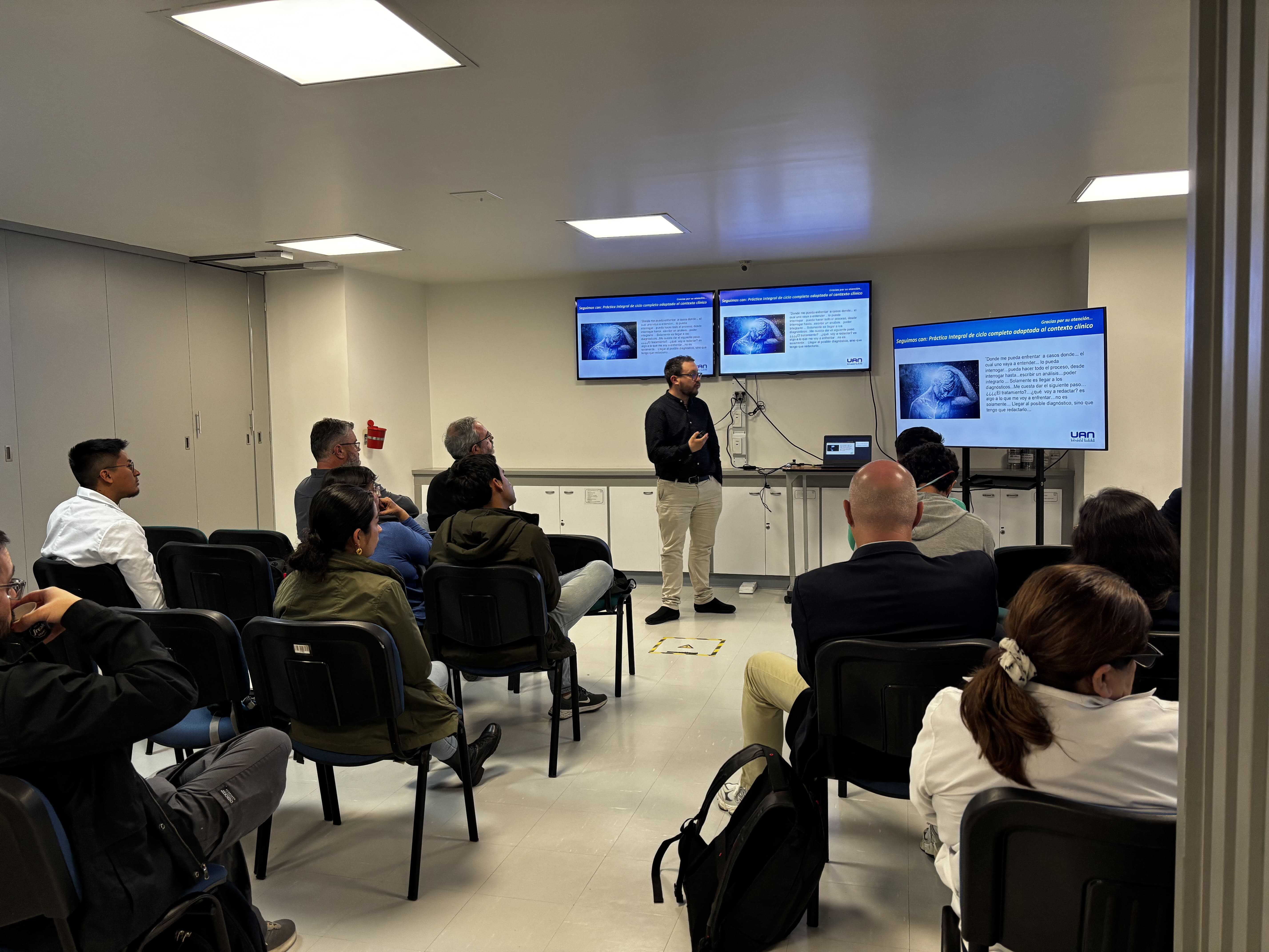 Un hombre imparte una presentación sobre temas médicos frente a un grupo de asistentes, apoyado en dos pantallas que muestran diapositivas informativas en el Centro de Simulación Clínica de la UAN